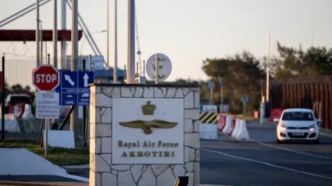 A landscape image of the entrance to RAF Akrotiri - a sign reads 'Royal Air Force Akrotiri' and a white car is in the background