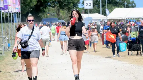 Festivalgoers arrive at the Isle of Wight Festival at Seaclose Park on June 19, 2025 in Newport, Isle of Wight two women can be seen walking along a dusty path with crowds behind them. one is drinking a bottle of water
