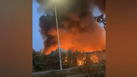 Georgie Collins Orange flames are trailing into the sky from a warehouse fire in Harwich, Essex. Followed by the flames is a plume of dark smoke rising up into the sky. 