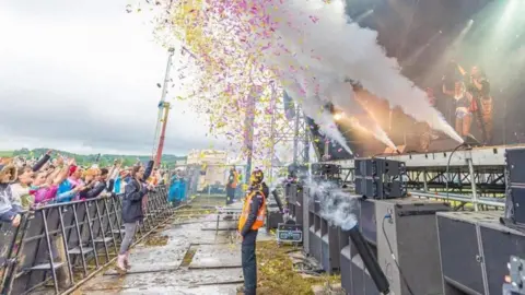 Kubix Kubix festival's stage photographed from the side. A security guard is standing in front of it as pink and yellow confetti flies in the air with stage smoke going off. Fans are lined behind a metal barrier and have their hands up in the air.