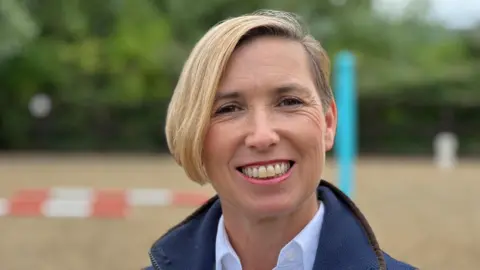 A middle aged woman with short blonde hair which is longer on one side stands in an outdoor riding arena, smiling. There is a show jump behind her. She is wearing a blue fleece with brown piping on top of a white shirt.