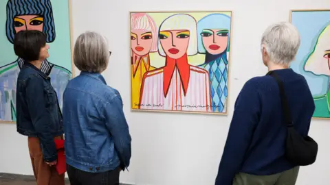 PA Media Three women look on at paintings on the wall of a gallery. The central painting depicts three women in colourful clothing in a 2D style, with portraits of solo women in a similar style either side.