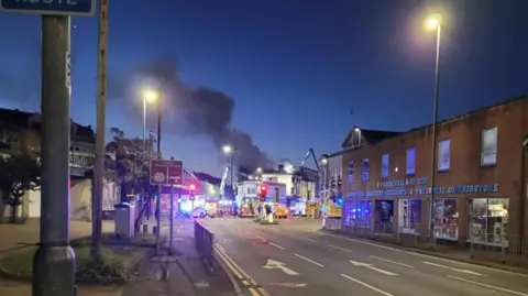 Liam Martin Thick smoke can be seen rising from a large building at the end of a three lane road. There are multiple emergency vehicles gathered around the site, with blue lights reflecting off shop windows.