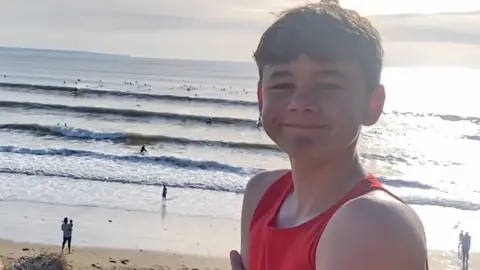 Neil Phillips Oscar is smiling at the camera as he stands with Saunton Sands stretching out behind him on a sunny day. The beach and sea are busy with surfers, swimmers and walkers.