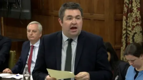 MP Shaun Davies addressing a debate in parliament's Grand Committee Room, holding notes, wearing a navy suit, white shirt and green tie