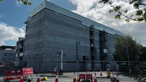The picture shows a large multi storey building completely wrapped in grey protective sheeting, with scaffolding visible along the right hand side where work appears to be ongoing. The wrapping covers almost the entire structure, leaving only small gaps where scaffolding platforms and metal poles can be seen. The building sits on a street corner, and the road in front is shut off with metal barriers, red and white cones and temporary fencing. A red sign in the foreground reads “Road closed"