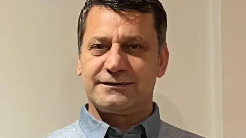 A man with short dark hair and wearing a light blue shirt stands in front of a pale pink wall