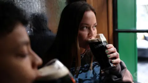 Reuters A woman with dark hair drinks a pint of Guinness beer in a pub while looking out of green-lined window.