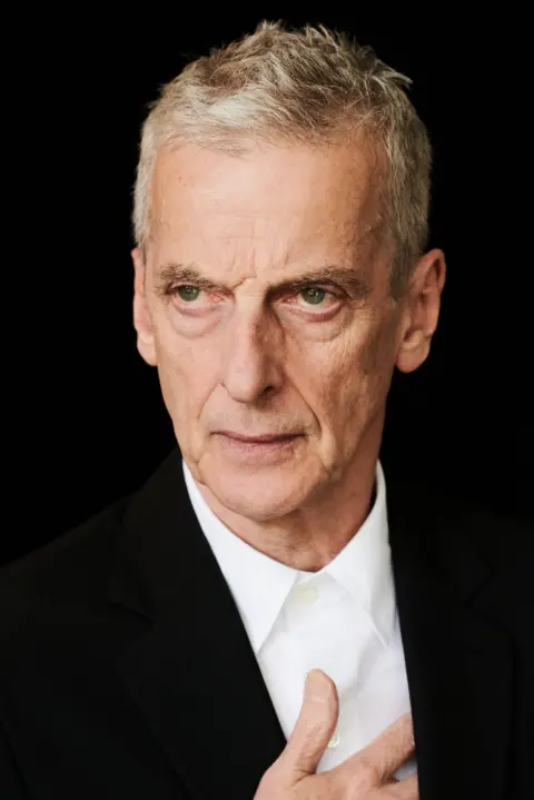 Last Night From Glasgow Peter Capaldi looking slightly off camera. He is wearing a black suit and white shirt and has his right hand to his heart