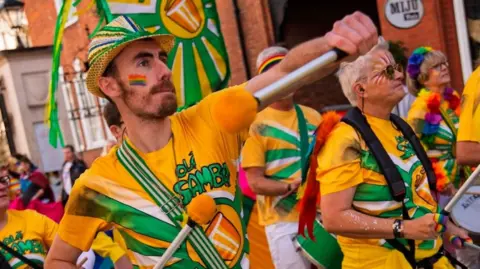 Pride in Gloucestershire 2025 Samba drummers wearing yellow and green costumes as part of the Pride in Gloucestershire march in 2025. The drummer in the foreground has a rainbow design painted on his cheek.