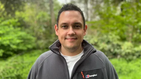 Jamie Andersen, who has cropped black hair, dark coloured eyes and is clean shaven, wearing a dark grey zip-up fleece and white t-shirt underneath, looking into camera with trees in the background