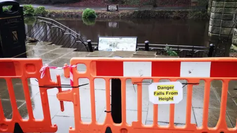 A wet, rain‑soaked walkway that has been blocked off by bright orange safety barriers. A yellow sign attached to the barrier reads “Danger Falls From Height", warning people not to proceed. Behind the barrier, a damaged railing can be seen bending down toward a brown, swollen river, suggesting that recent flooding or erosion may have caused the collapse. On the left side of the image, two black litter bins stand near the edge of the path, while on the right, a stone structure rises out of the frame.