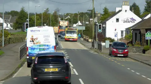 David Dixon Multiple cars, a van, a bus and a lorry travelling through the centre Springholm village on the A75. A restaurant and housing is shown on the side of the road and a set of traffic lights are positioned half way up the road.