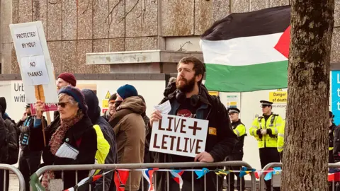 A picture of a number of flags and banners on a silver barrier. There is a number of people stood behind the barrier. There is a man holding a 'LIVE+LETLIVE'