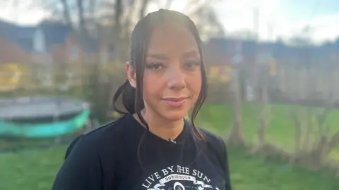 A woman wearing a black T-shirt stands in her garden looking at the camera. The background is bright and sunny but a bit blurred. 