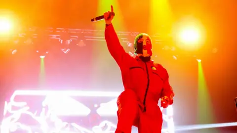 Getty Images A member of Kneecap see on stage at Coachella 2025. There are red strobe lights around him and he is wearing a red boiler suit with a balaclava featuring the Irish flag.