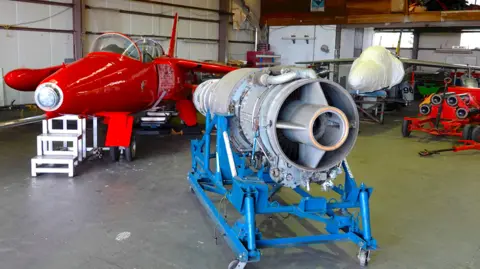 The mark 101 Orpheus engine supported by a blue rig. It is placed in a warehouse and stands next to a red Gnat aircraft which belongs to Red Gnat limited. Another small aircraft can be seen in the background but is covered in a white tarpaulin.