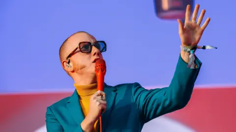 BBC The lead singer of Two Door Cinema Club, Alex Trimble, performing holding a red microphone and wearing a blue suit