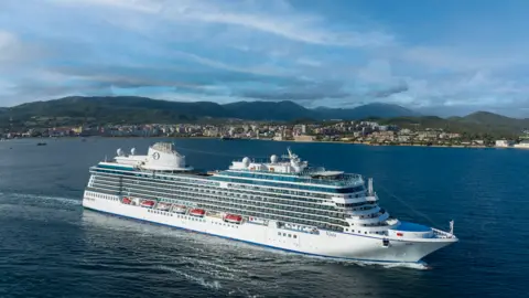 Oceania A large white luxury cruise ship is seen in the water with mountains and area of housing on the land behind it.