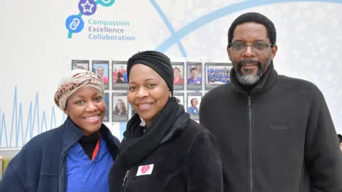 Royal Papworth Hospital NHS Foundation Trust Ms Mushonga's daughter is smiling at the camera while standing next to her mum on the left. She has brown eyes and is wearing a blue fleece. Ms Mushonga's husband is standing to her right. He is wearing glasses and has black hair and black and grey facial hair.