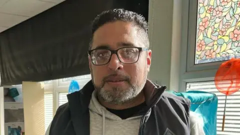 A man wearing a grey jumper and a navy gilet stood in a community centre. he wears black-rimmed glasses and has tight curled black hair. Behind him are windows with blinds on them, a coat rail and a white table.