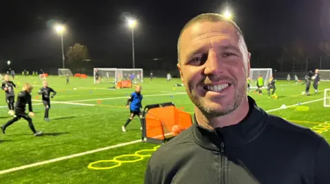 Tommy Coyle smiles into the camera. He has short, shaved hair and a stubble beard. He is standing at the side of a football pitch bathed in floodlights. A number of children are visible in the background playing football.