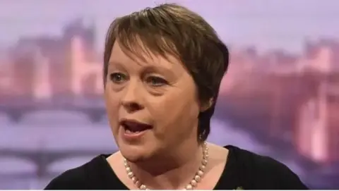 Maria Eagle appearing in a BBC television studio. She has brown choppy hair to her shoulders and is wearing a pearl necklace. She has a black top on. 