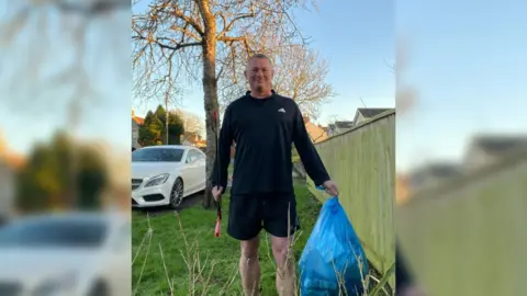 A man wearing a black jumper and shorts stands on a patch of grass with a litter-picking device and blue waste bag.