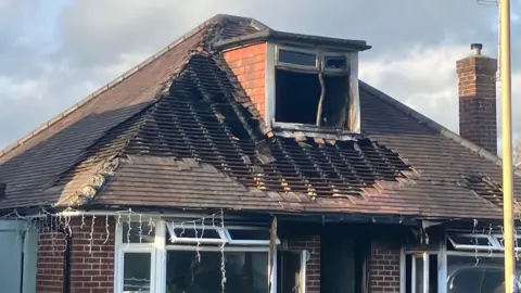 A shot of a traditional brick house with a terraced roof which has been significantly damaged by fire. Many of the roof's tiles are completely missing and a box window in the attic has been blackened and warped. 