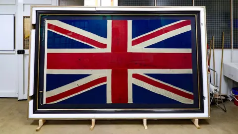 A large framed union jack flag is displayed upright indoors, leaning on wooden supports in what seems to be a workshop or conservation space. The flag shows visible creases, small marks, and wear. The red, white, and blue design fills most of the frame while tools, and equipment are visible in the background.