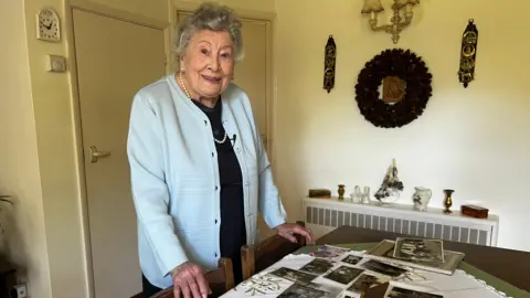 Silvia Verrall looks at the camera and smiles, she is standing next to a table covered in old photographs and is wearing a pale blue jacket