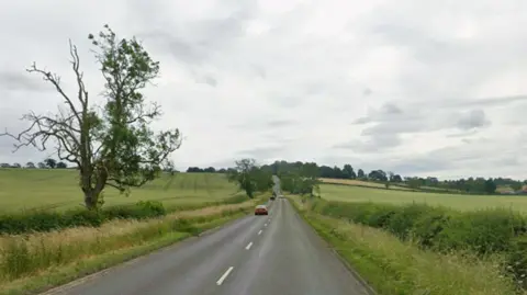 A Google streetview of the A6003 Oakham Road. The road is surrounded by green fields. The rear of a red car can be seen in the distance.