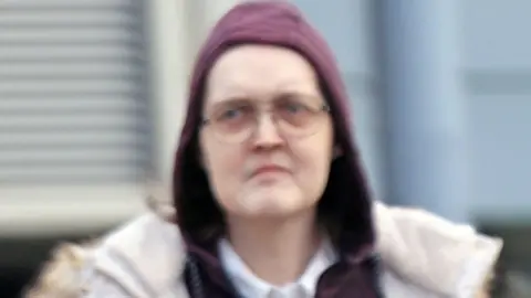Vic Rodrick A transgender woman, with a stern expression wearing glasses and a burgundy hoodie which is covering her head.