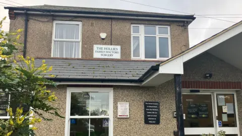 The front of The Hollies Surgery, which is a beige-coloured two-storey pebble-dash house. A sign reads THE HOLLIES FAMILY DOCTORS SURGERY.