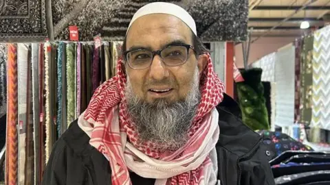 Ibrahim has a white cap, red and white chequered scarf and a balck anorak. Hre has a grey beard and black spectacles and stands in front of textilew display.