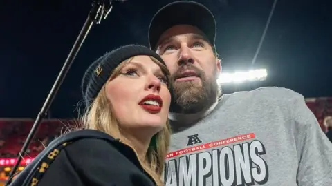 Getty Images Taylor Swift stands next to her fiancé Travis Kelce in an American football stadium. She wears a black hooded sweatshirt with a black hat. It wears a gray sweater with the word 