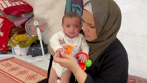 Woman with a veil holds baby girl who looks distressed and is crying inside a tent in Gaza. There is a fan and some blankets behind them in the background.
