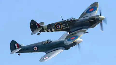 Longleat A pair of BBMF Spitfires flying side by side in blue sky with propellers and silver-grey bodies and the iconic blue, red and white bullseye insignia painted on their wings.