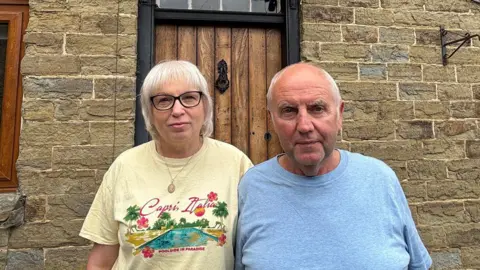 A man and a woman are standing in front of a brick cottage. The woman has short white blonde hair and black glasses. She is wearing a yellow t-shirt. The man has very short white hair and is wearing a light blue grey t-shirt