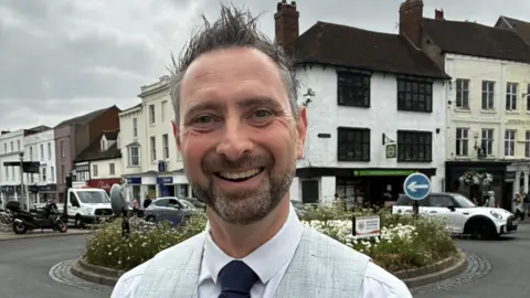 Aaron has spiky grey hair and is wearing a white shirt, grey waistcoat and a black tie. He stands in front of a roundabout decorated with greenery and flowers. There are wite buildings in the background and cars driving past them