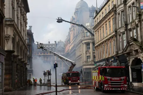 PA Media Fire engines at the site of the Union Corner fire in Glasgow. Firefighters use two large hydraulic lifts to aim water from hoses into the smoking building. It is a built up area with a street lined with buildings several floors high.