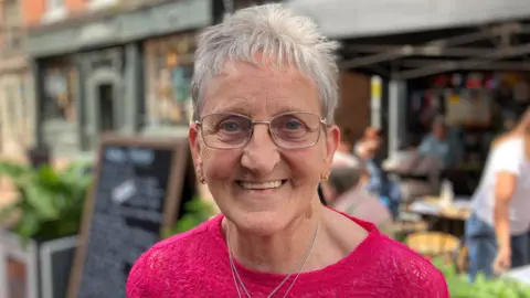 A woman with short grey hair and glasses, wearing a pink top, smiles at the camera