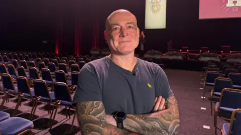 BBC/Eleanor Doyle Dr Beardon has his arms crossed and smiles at the camera as he stands in a hall with several rows of chairs in front of a stage. He has tattoos on both his arms and is wearing a grey t-shirt and watch on his left wrist.