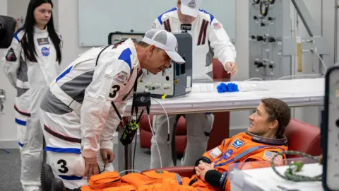 NASA Inside a bright white control room, a suited technician bends over an astronaut reclining in an orange launch-and-entry suit, checking equipment beside her. Other white-suited technicians stand and work at consoles and tables covered with cables. 