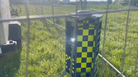 The three-foot hexagonal robot stands in a field next to the drilling unit inside a metal fence. It is covered in black and yellow checks.
