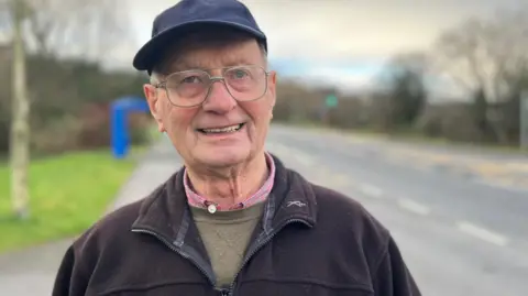 A man is smiling at the camera, he is wearing large silver-rimmed glasses. He has a dark blue hat on and is wearing a black zipped jumper, underneath he has a grey jumper and you can see the collars of a pink shirt peaking through. He is stood on the roadside and behind him is a blurry image of a grass verge to the left and road on the right. The man is the focus of the picture.