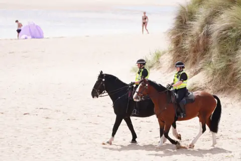 PA Media Two mounted police officers on horses next to sand dunes on Balmedie Beach ahead of a visit by the US President Donald Trump and Prime Minister Sir Keir Starmer to the president's Menie golf course in Aberdeenshire. Two people are on the beach in the background - a woman in a bikini and a boy at a sun shelter.
