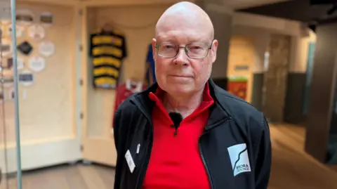 Dr Nick Lindsay is standing inside Brora Heritage Centre. He has a bald head and is wearing glasses, and a black fleece over a red top. 