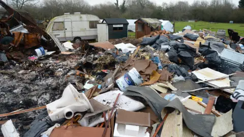 Large amounts of rubbish lies in a field. A large part of the pile has been burned but a toilet, tins of paint, carpet and wood are visible. The charred remains of some of the waste can be seen on the left of the image after it has been burned. A small caravan and two small sheds are visible in the background.