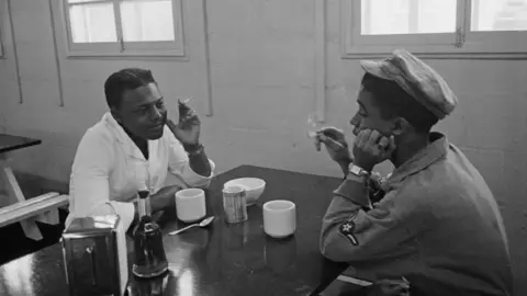 Haywood Magee/Getty Images Two people are sitting opposite each other in a canteen. There is a table between them and crockery and other items are on it. One person is wearing a white overall and the other is in a military uniform.